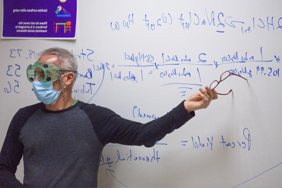 A faculty member, wearing safety goggles and a face mask, points at a formula on a whiteboard.